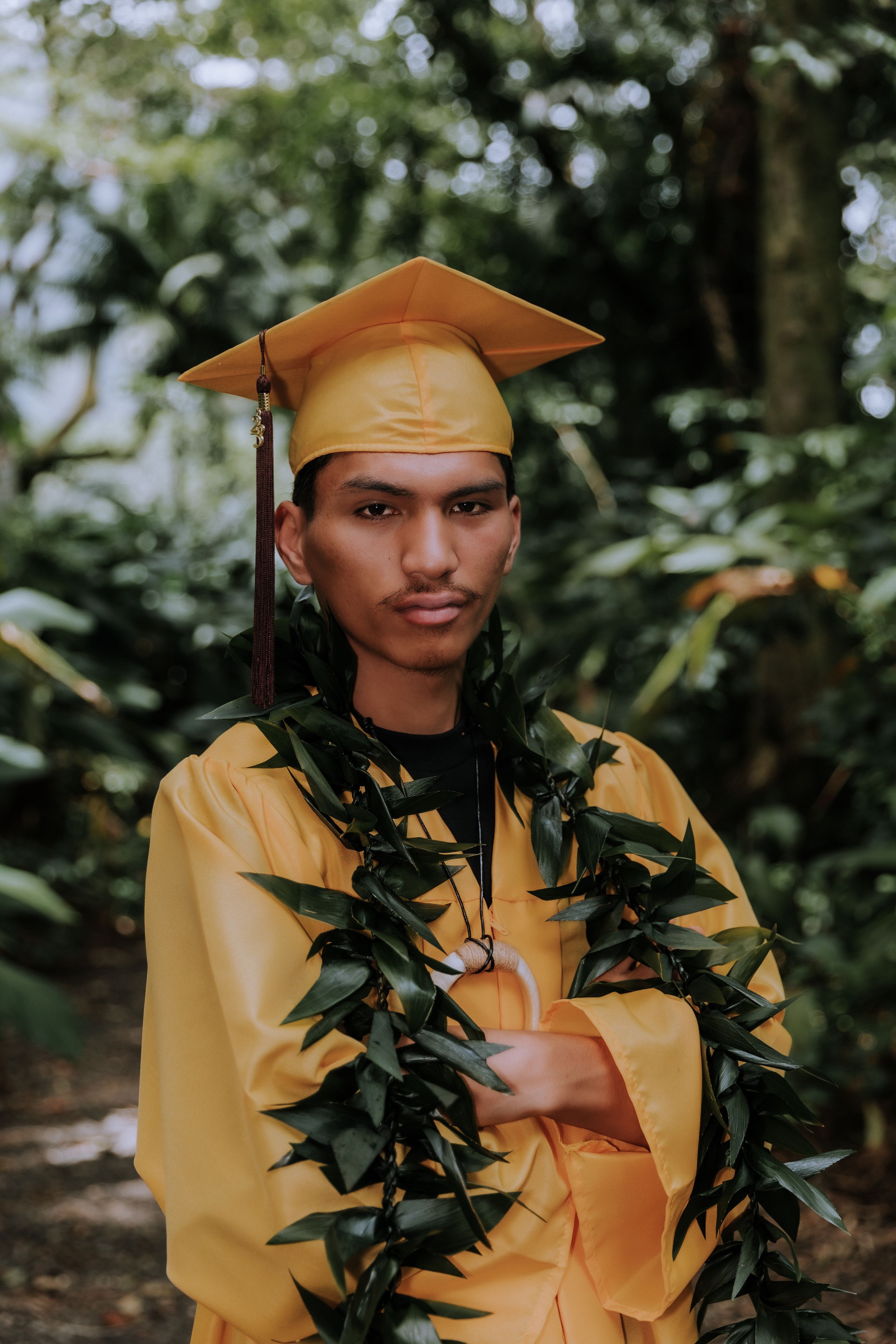 Graduate in gold cap and gown with green leaf lei in garden setting.
