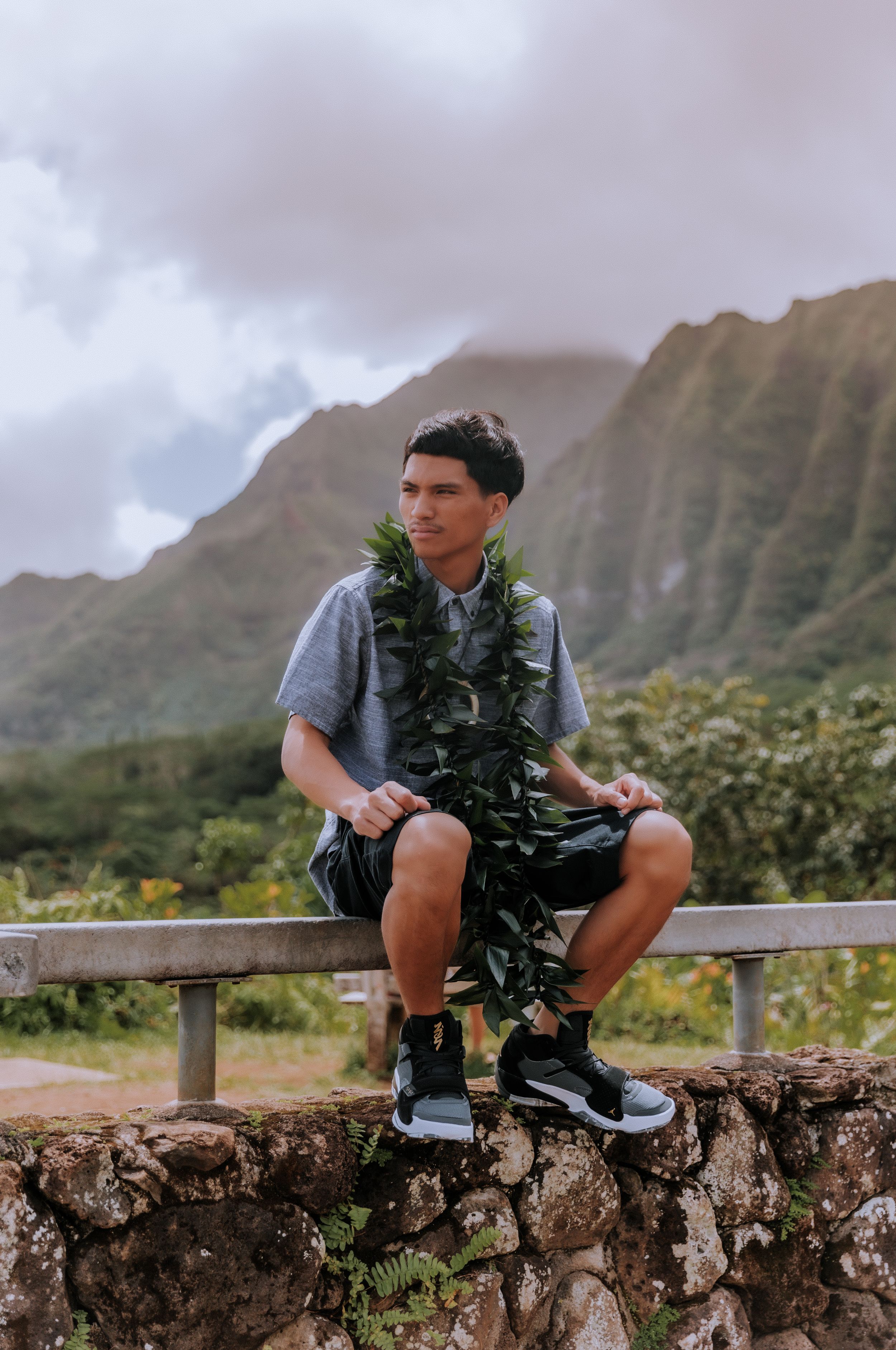 Person wearing dark lei sits on stone wall with lush mountain landscape in background.
