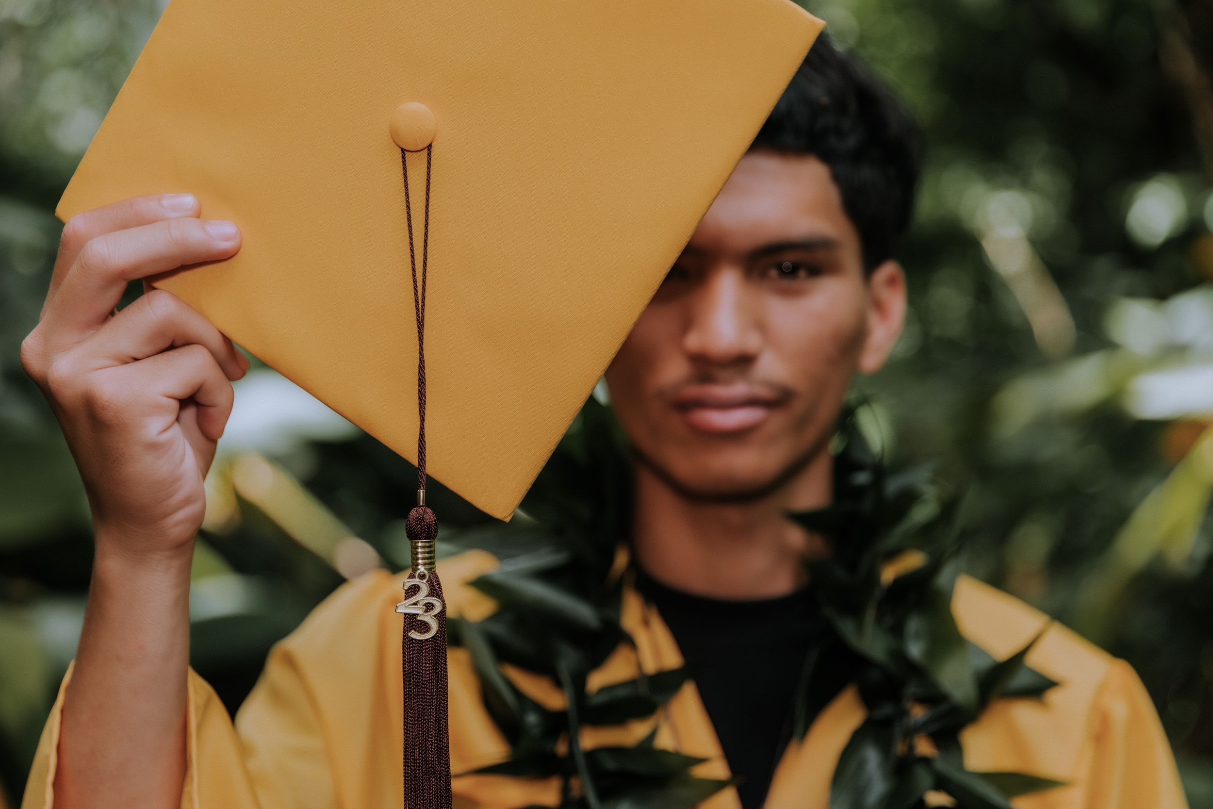 Gold graduation cap held up against green foliage background.