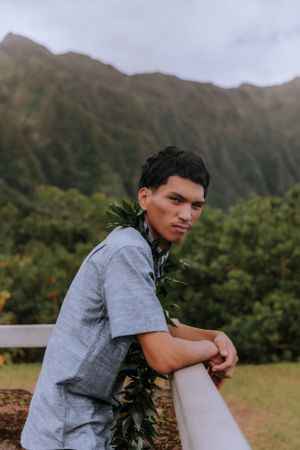 Individual in grey shirt with traditional lei sits outdoors with mountain scenery.