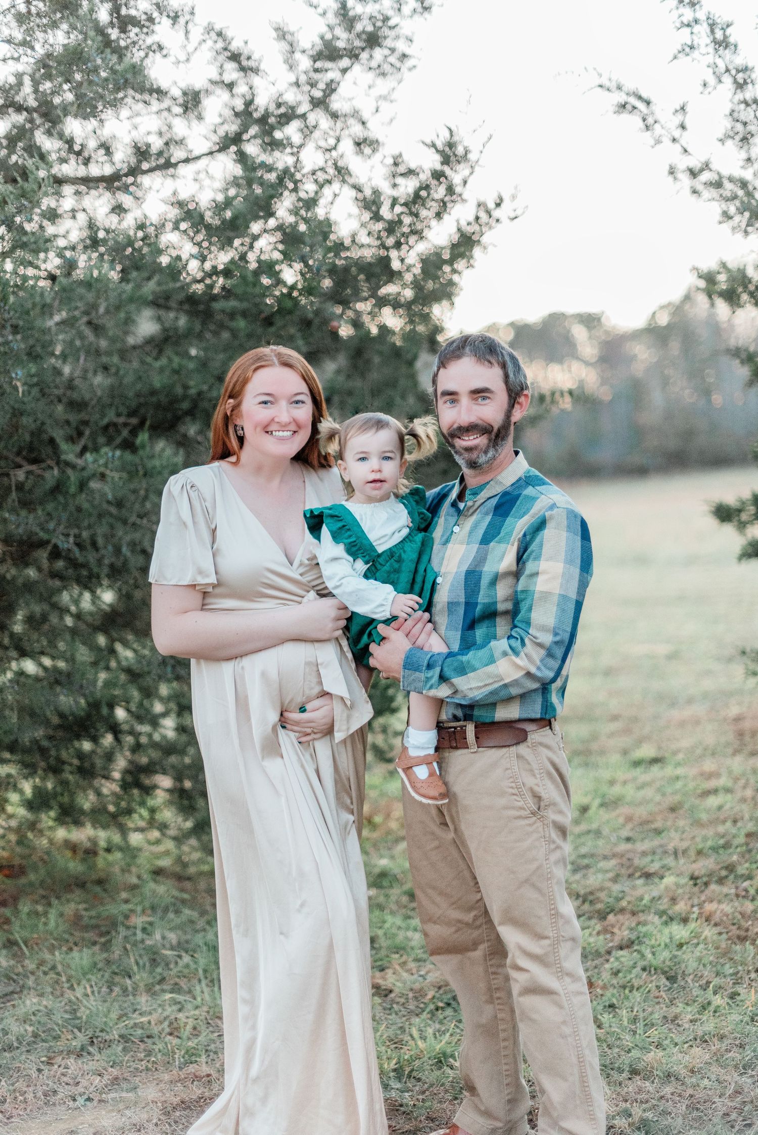 A family poses together outdoors in a natural setting with trees and grass at sunset.