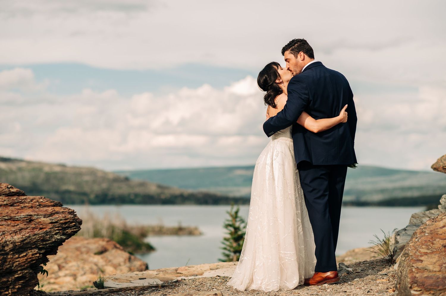 couple in wedding attire kissing with the sun shining on them in glacier national park