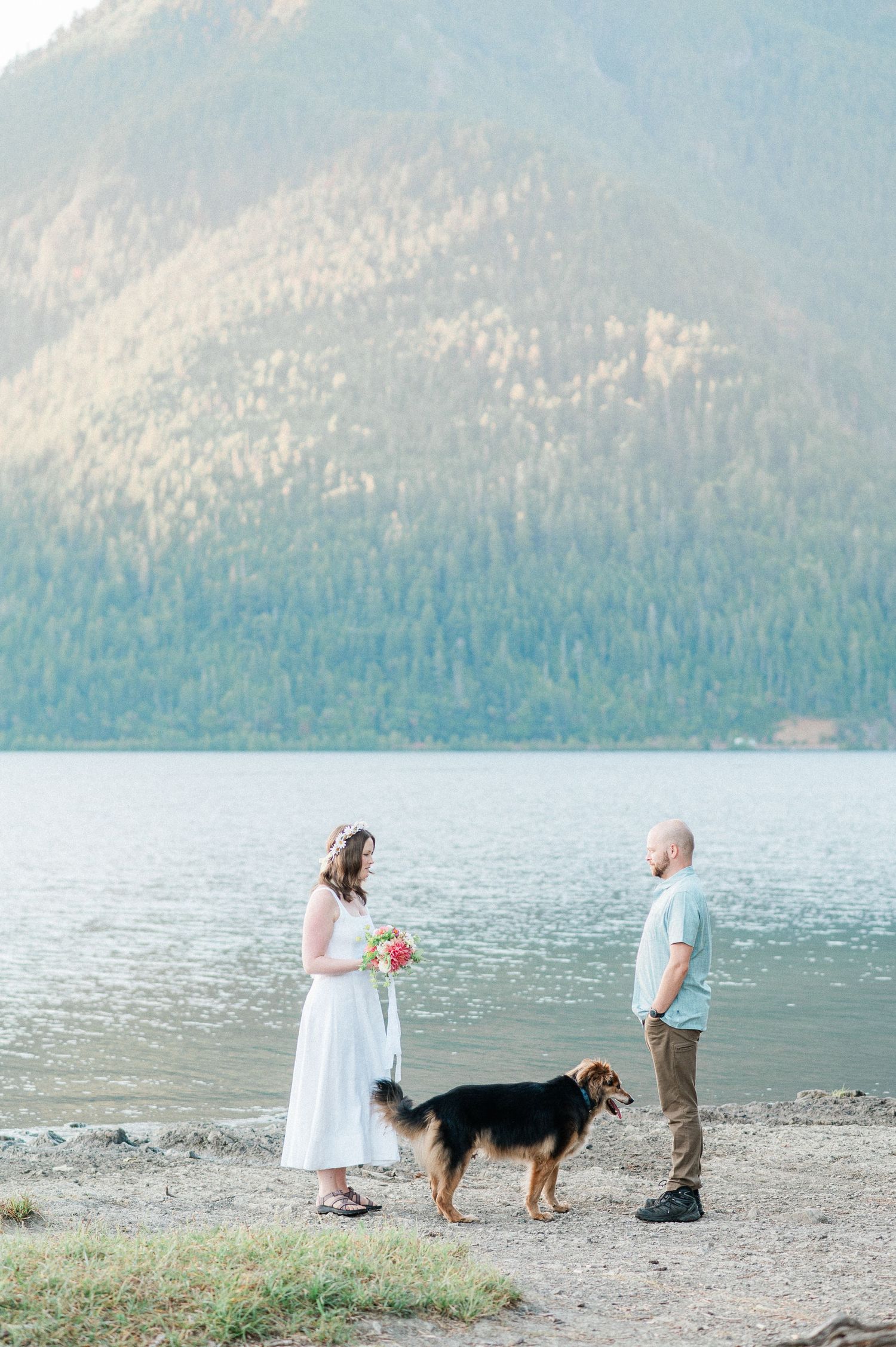 A couple and their dog walk along a scenic mountain lake shore on a peaceful summer day at Lake Crescent. Photo by Pine + Vow.