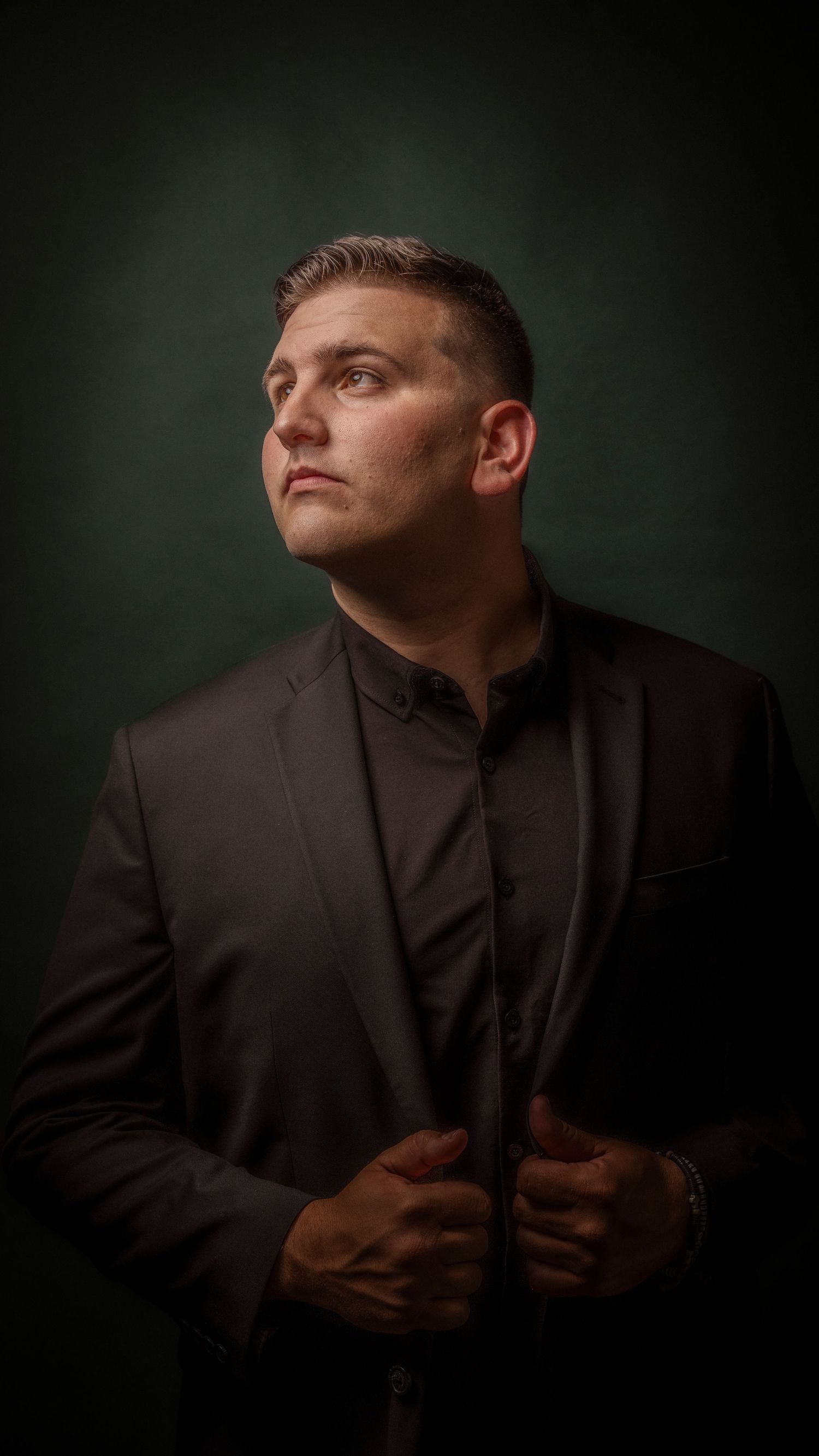 Confident man in black suit jacket posing against dark green studio background portrait.