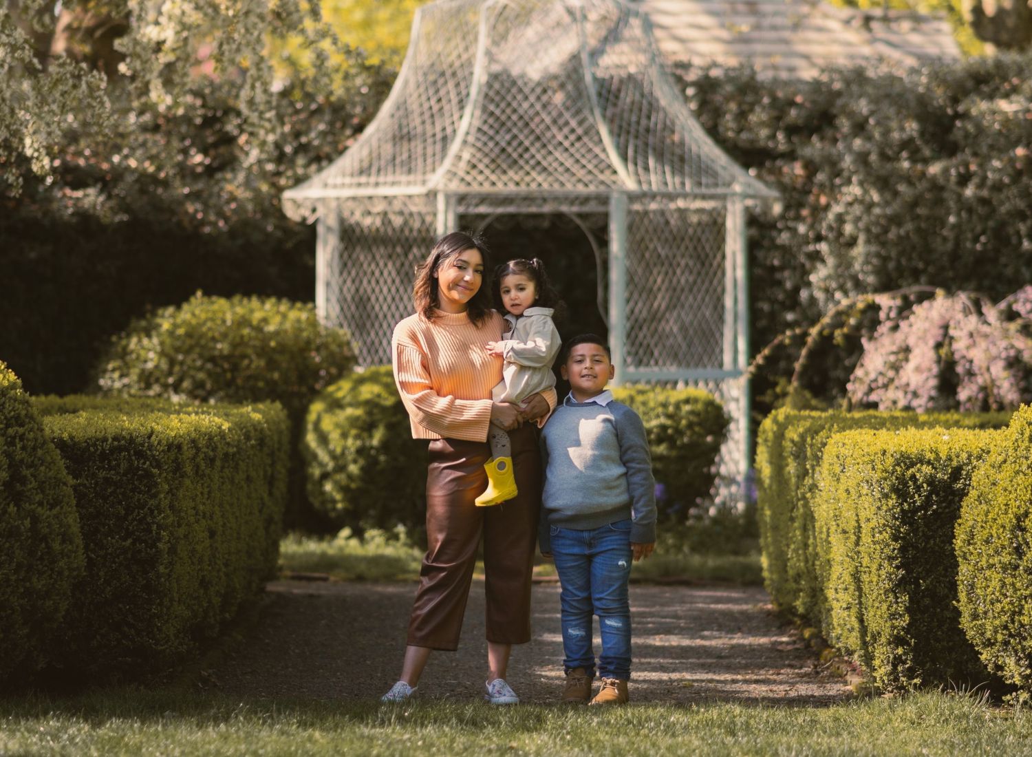 Mom holding daughter on hip with son hugging her on the right. Photo centered in front of cute pergola.