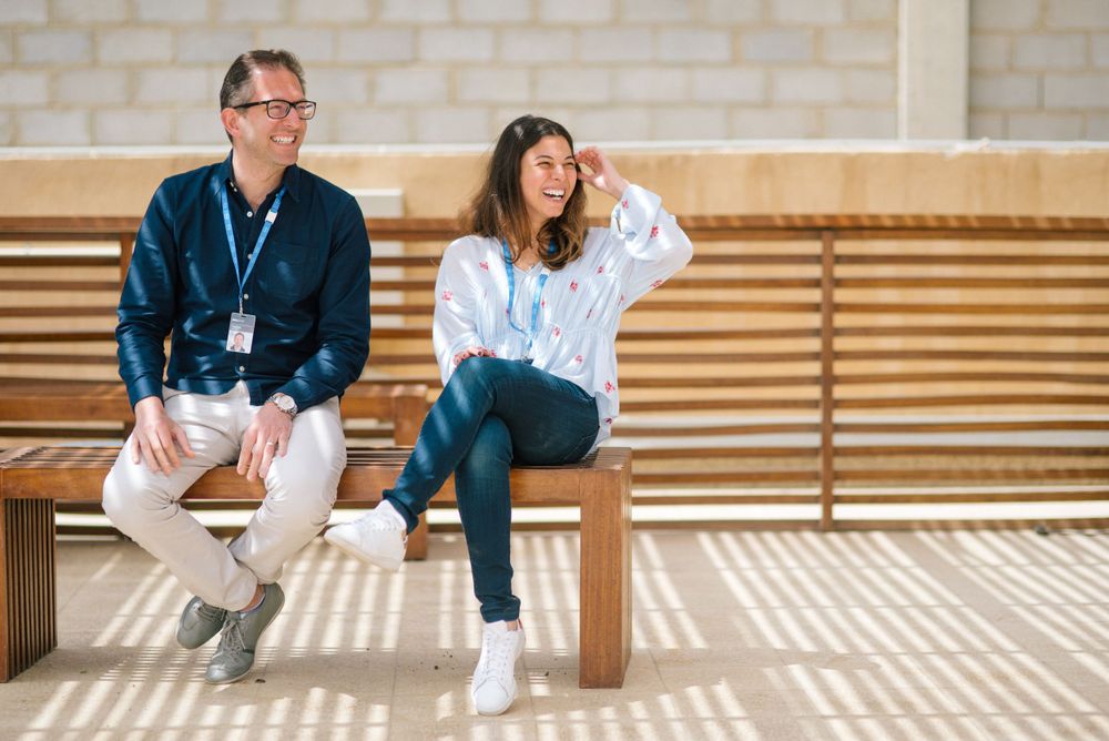 Two colleagues sitting on wooden bench in casual conversation during sunny day.