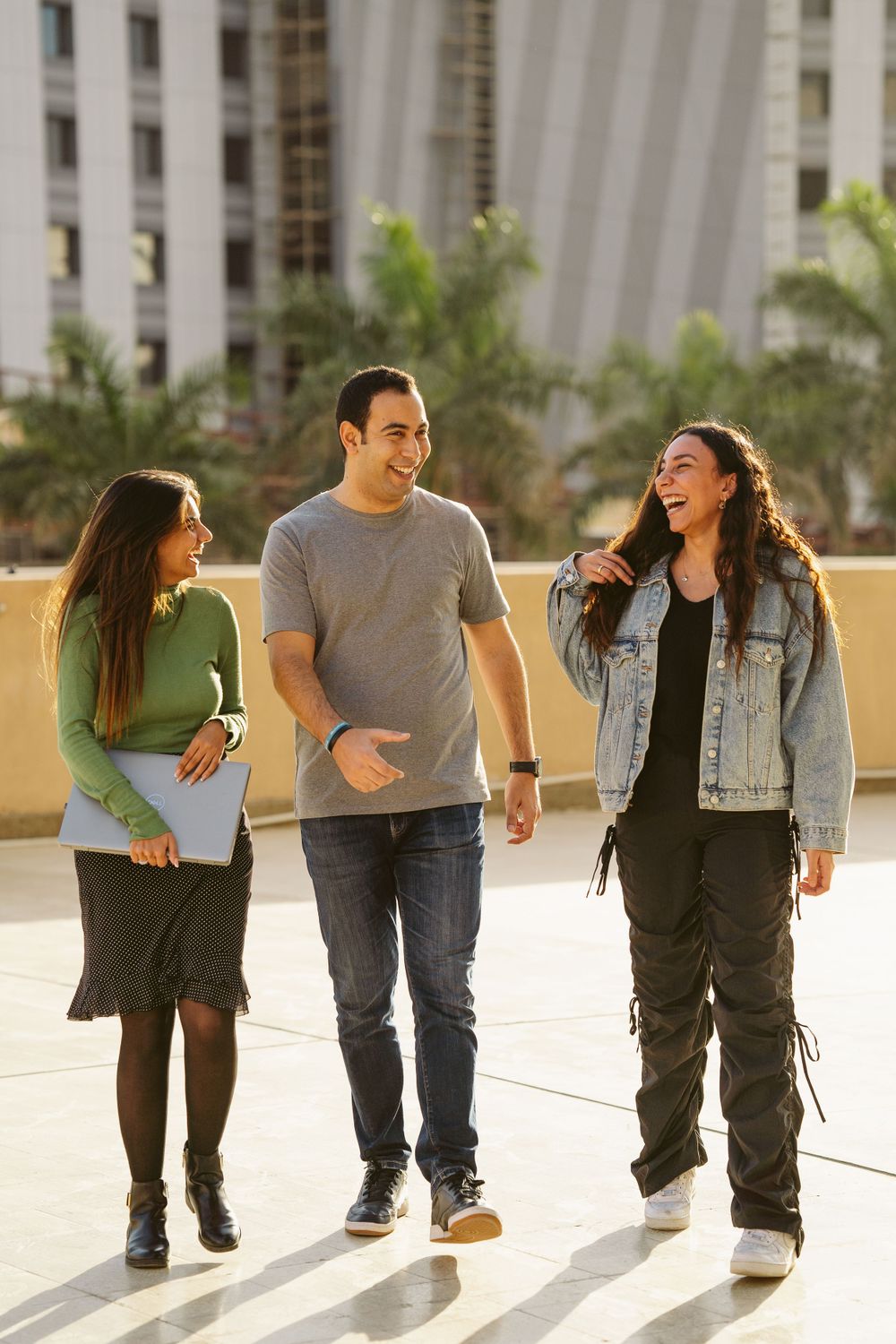 Three friends walking and laughing together on sunny campus pathway.
