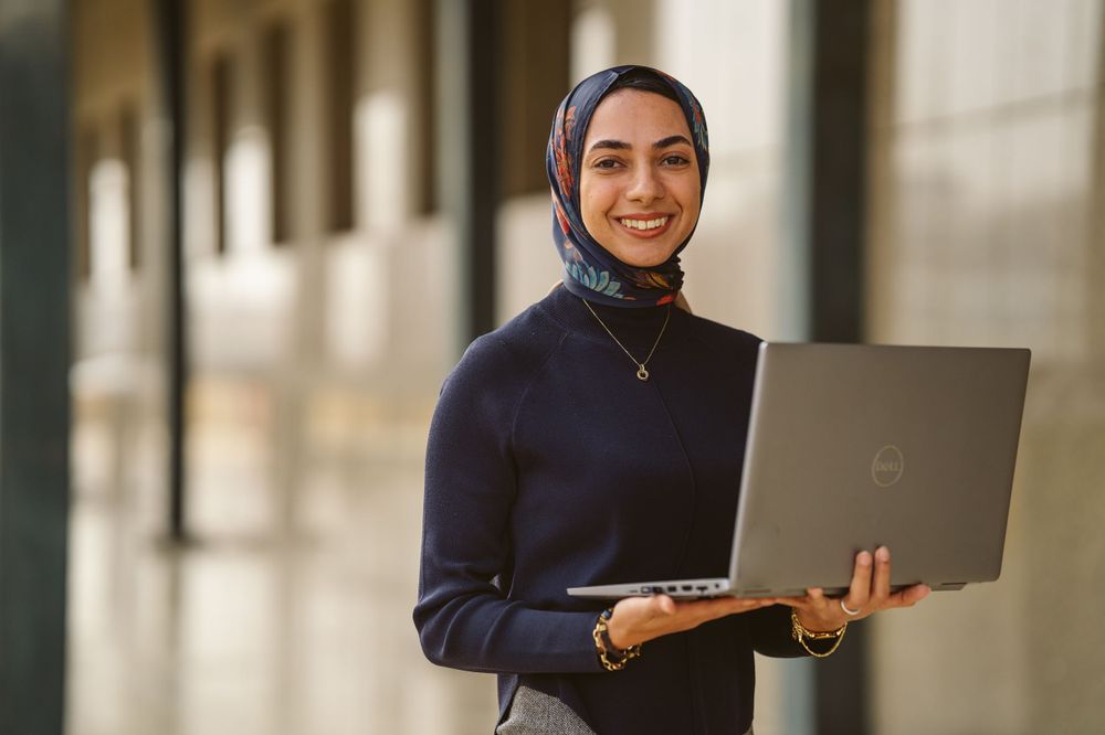 Professional woman in navy hijab smiling while working on laptop in bright hallway.