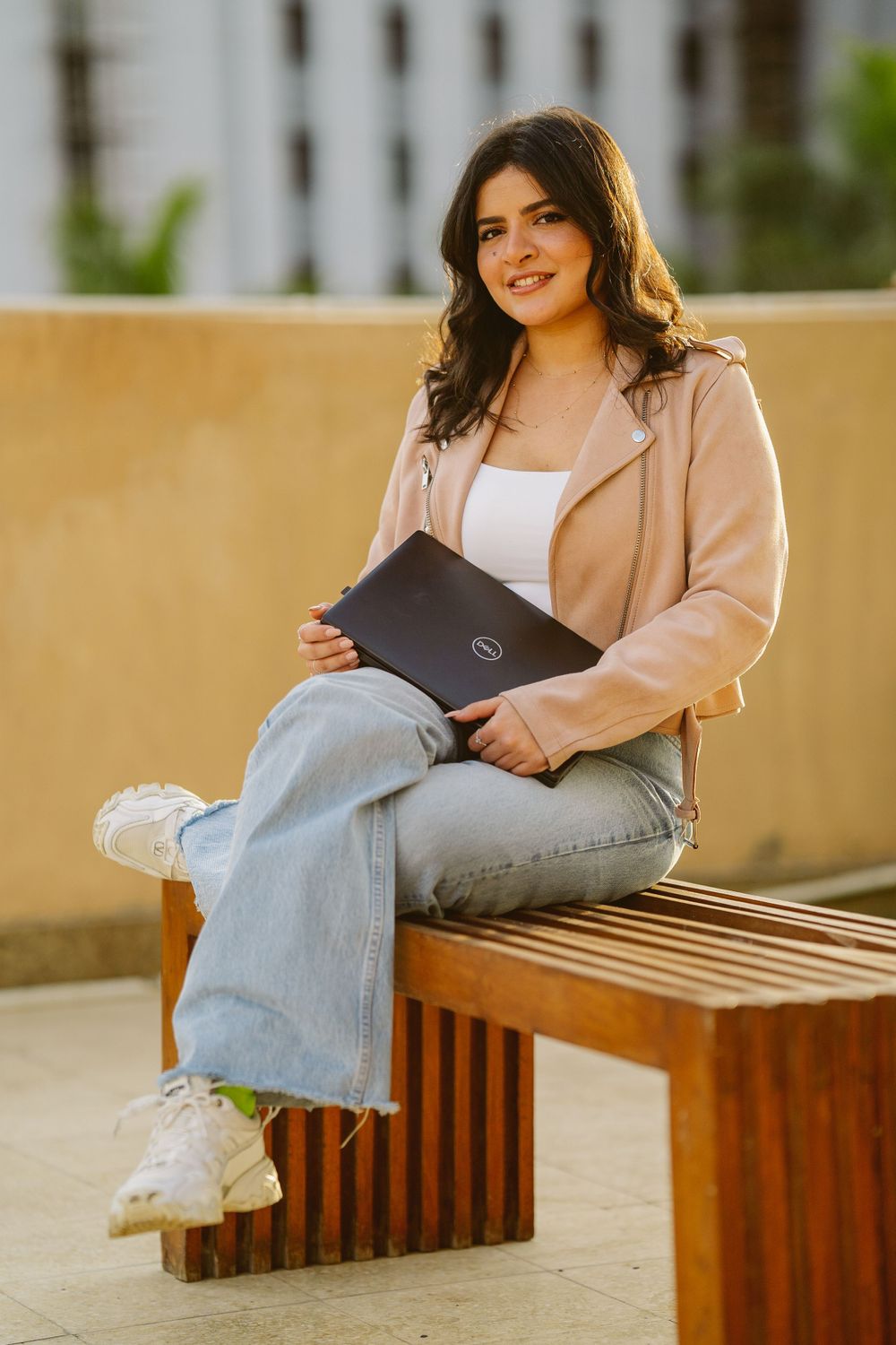 Student in beige jacket sitting on wooden bench with laptop outdoors.