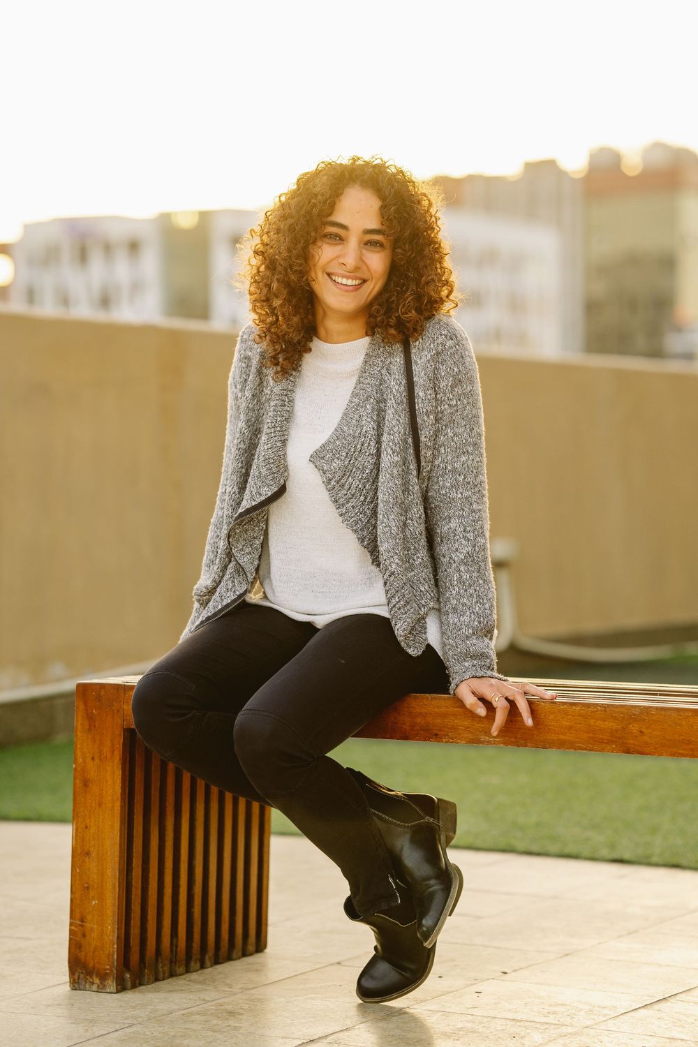 Person with curly hair sitting on wooden bench outdoors wearing grey cardigan and black pants.