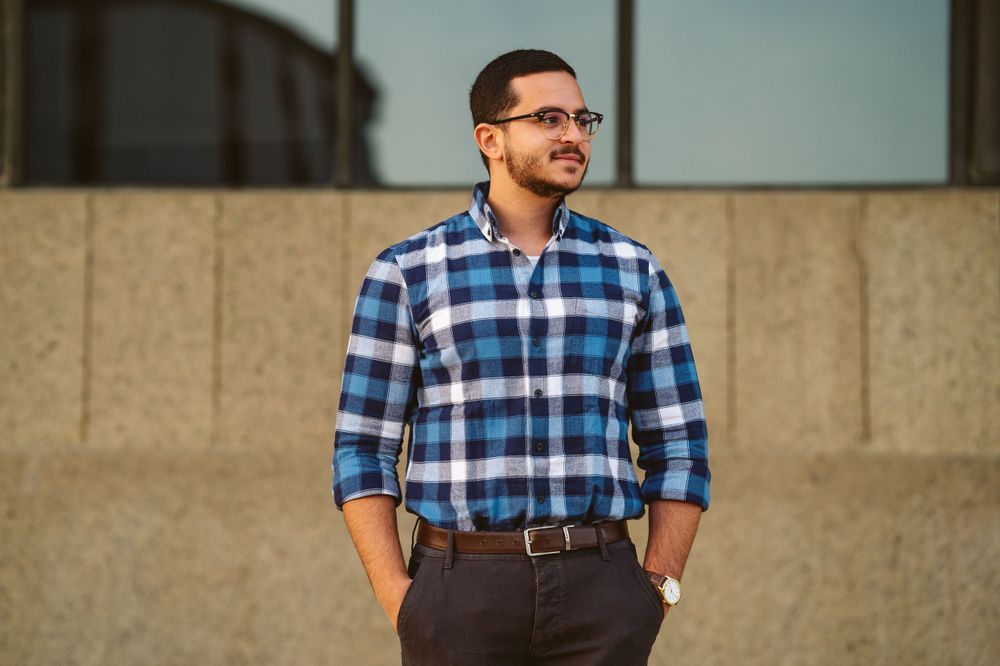 Professional in plaid button-down shirt and glasses standing against concrete wall at sunset.