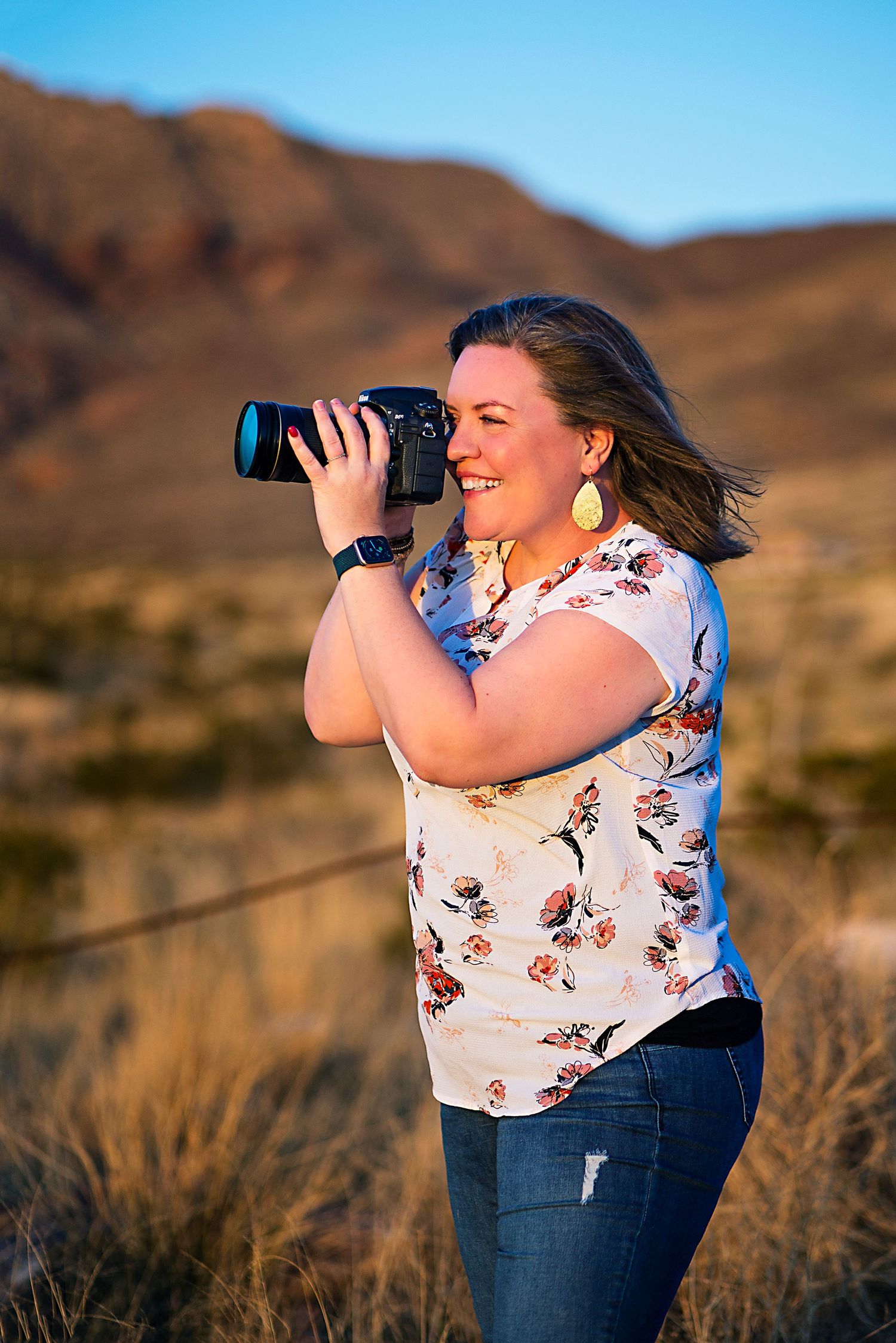 A photographer in a floral top captures photos in a desert landscape against brown mountains.