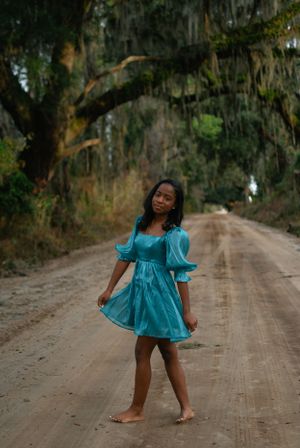 A person in a turquoise dress twirls playfully on a dirt road lined with Spanish moss-draped trees.