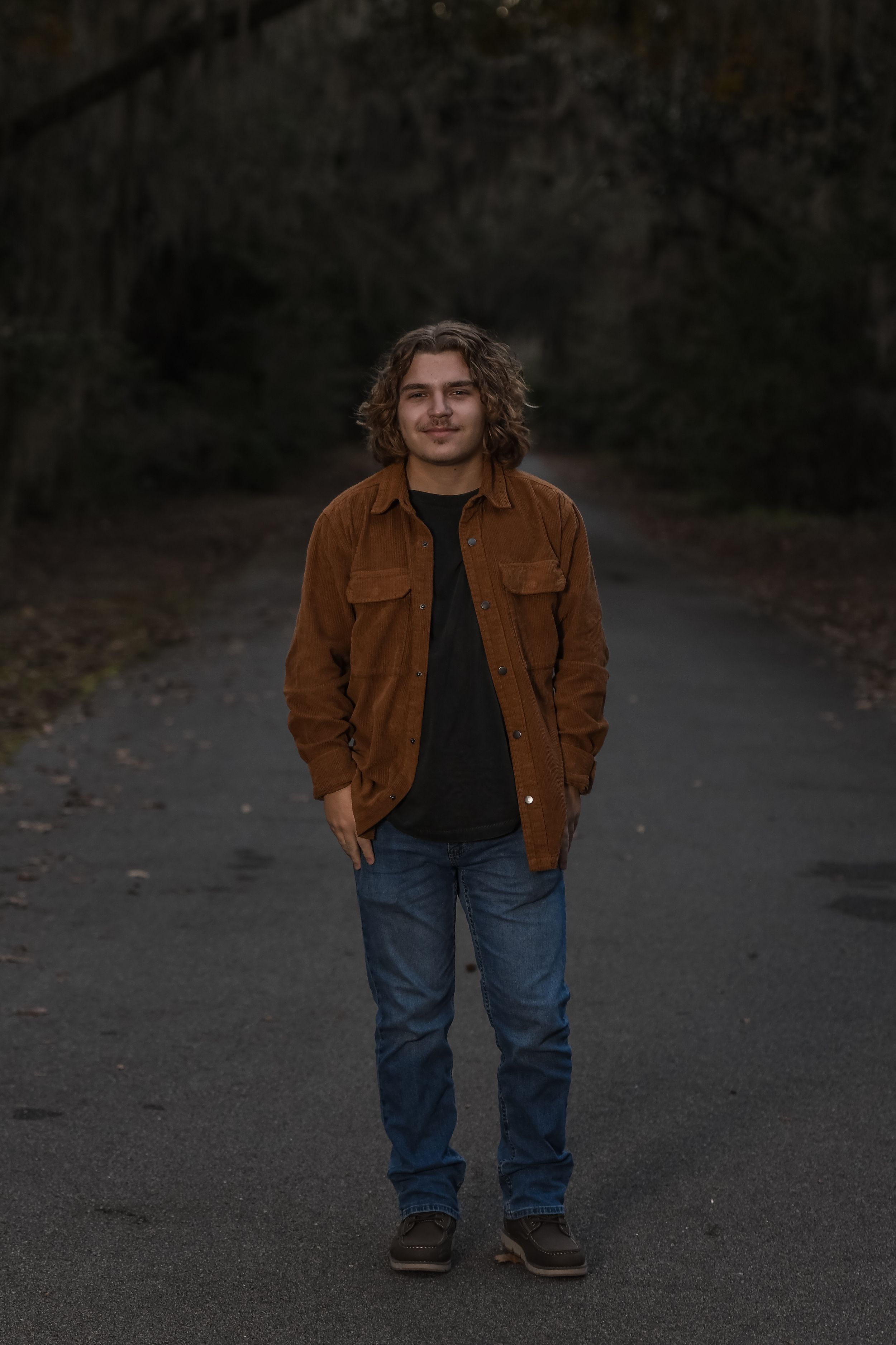 Person wearing brown corduroy jacket and jeans stands on forest path in moody lighting.