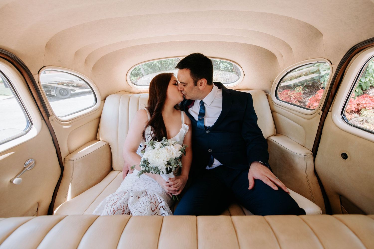 Romantic wedding couple sharing an intimate moment in the back seat of a vintage car with beige interior.