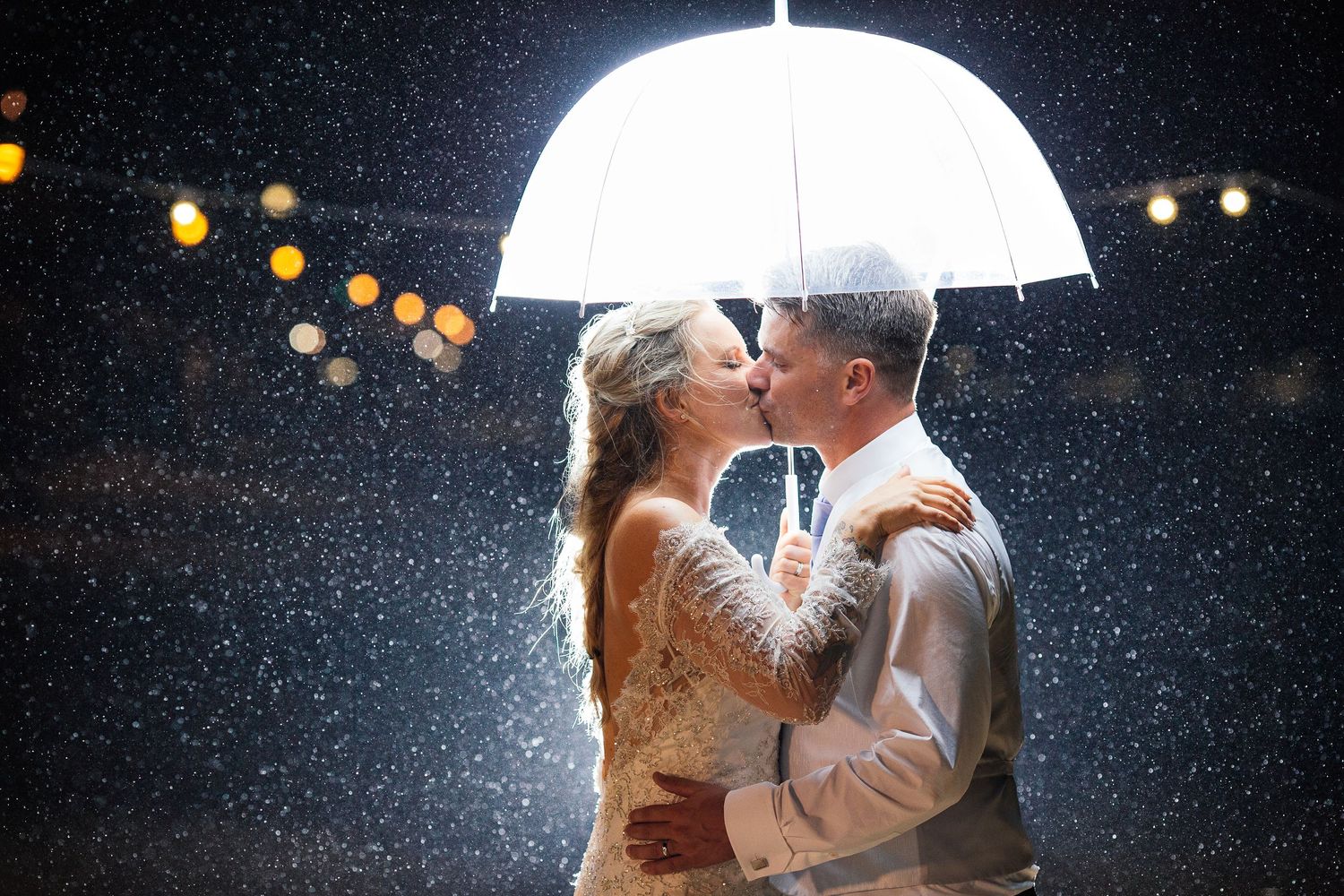 Romantic couple sharing a kiss under a white umbrella in the rain with glowing bokeh lights in the background.