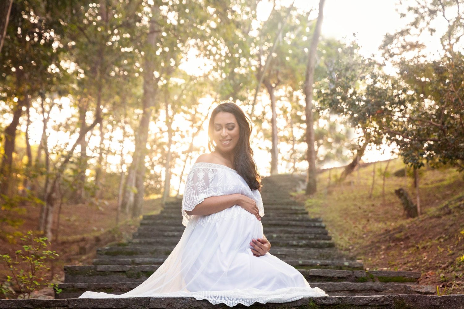 Elegant maternity photo of woman in white dress seated on outdoor stairs at sunset.