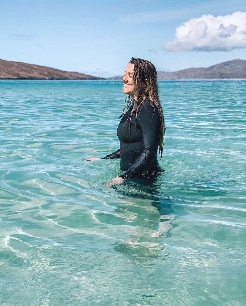 Woman in a black wetsuit standing in crystal clear turquoise ocean water with mountains in background.