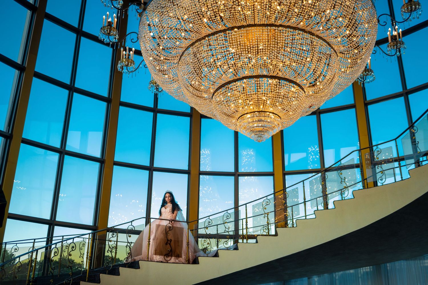 Dramatic portrait under grand crystal chandelier with floor-to-ceiling windows and curved architecture.