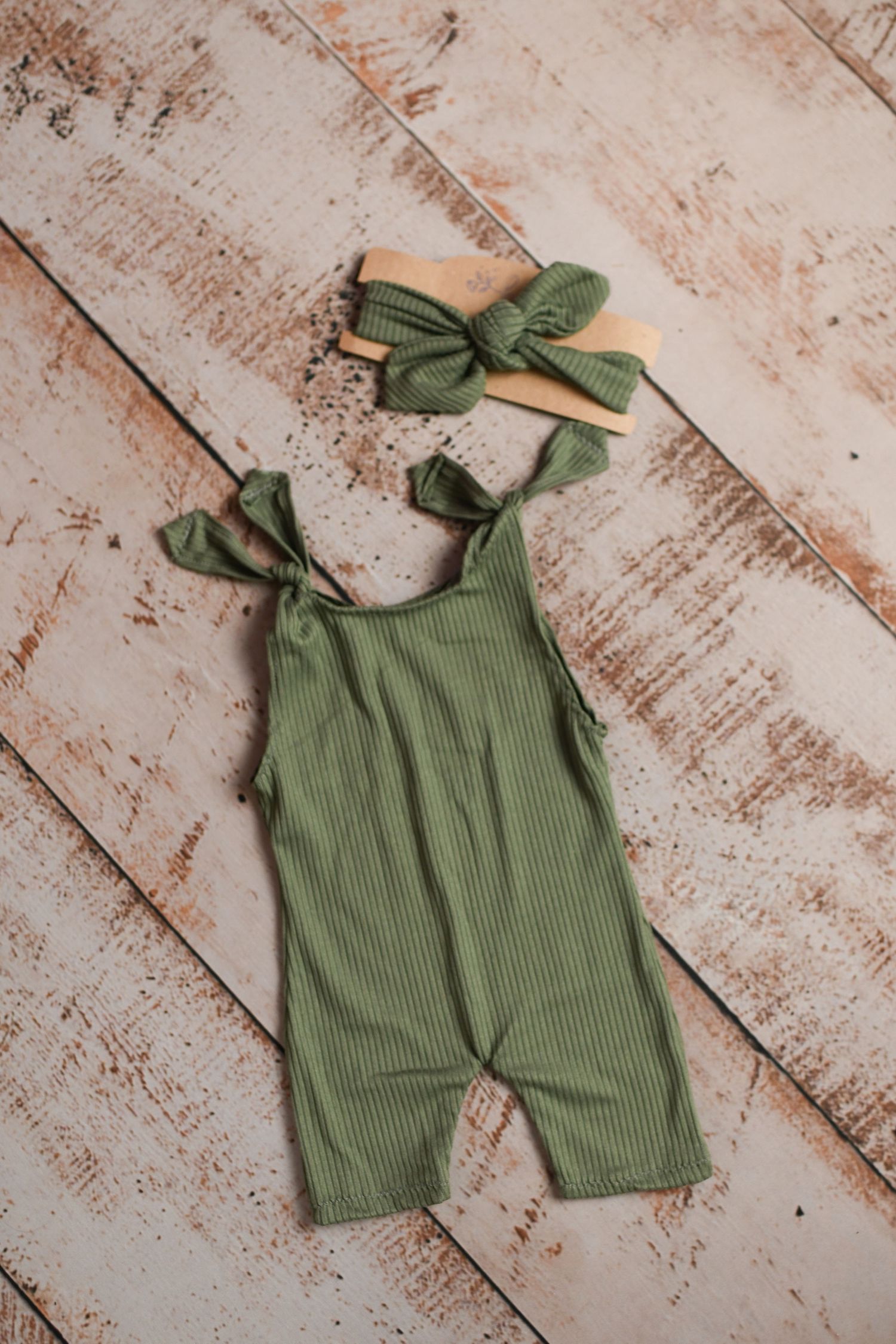 Olive green ribbed romper with bow straps displayed on rustic wooden backdrop.