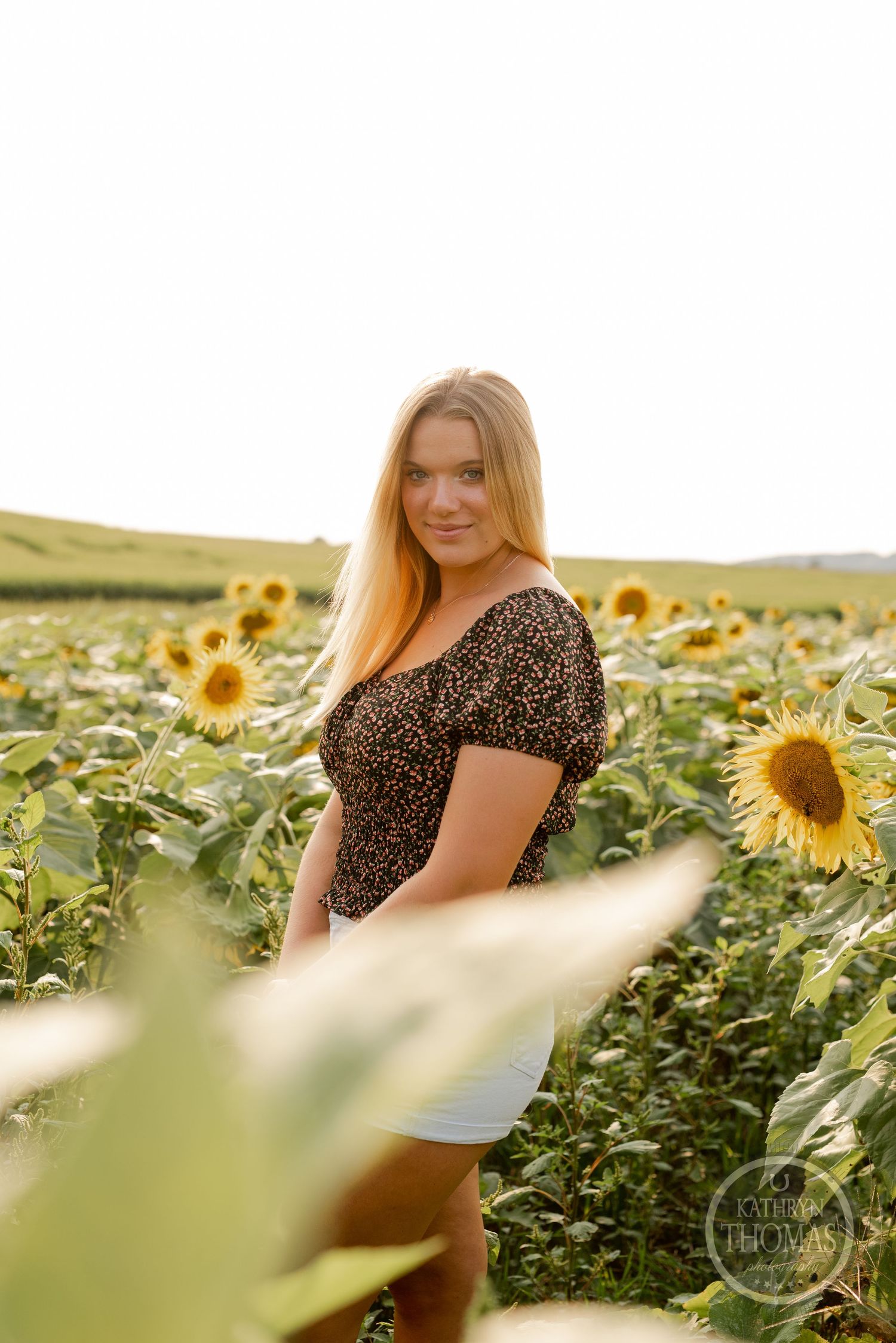 A candid shot in a sunflower field showing a person in a dark floral top against golden light.
