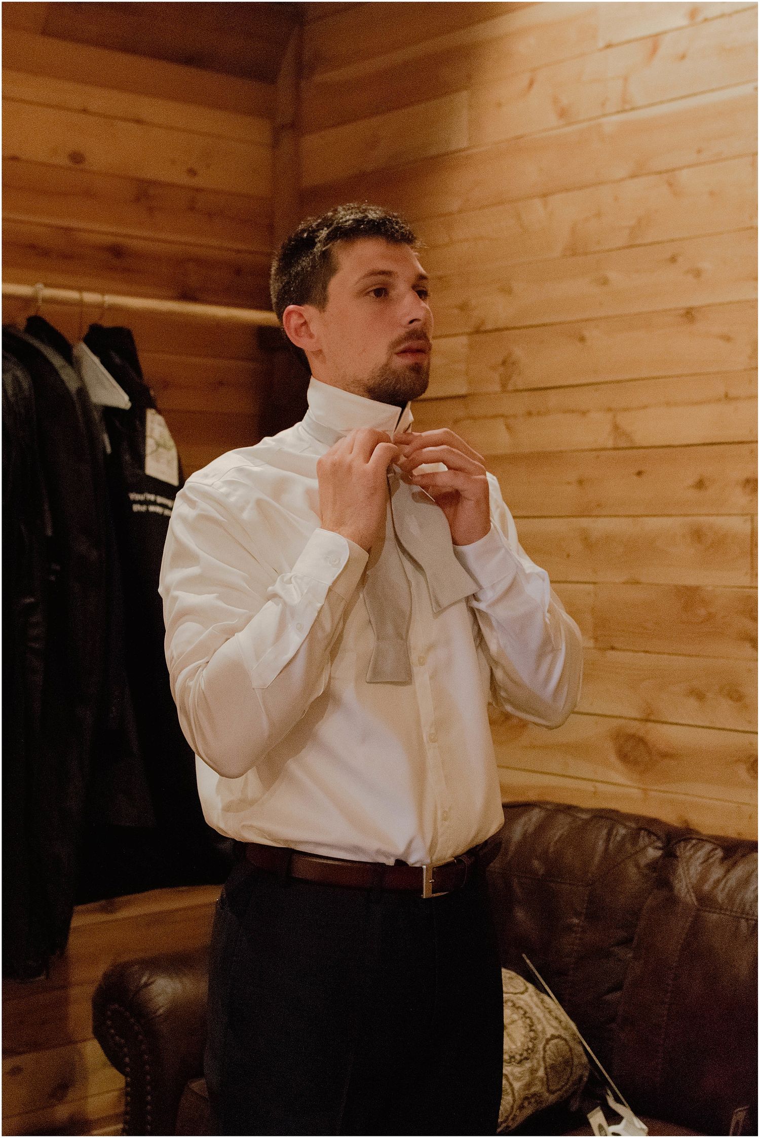 Groom adjusts his tie while getting ready in a wood-paneled room.