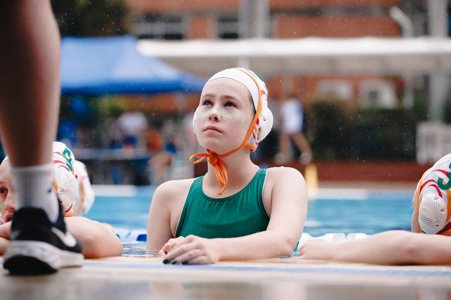 Water polo player in green suit and white cap resting at pool edge.