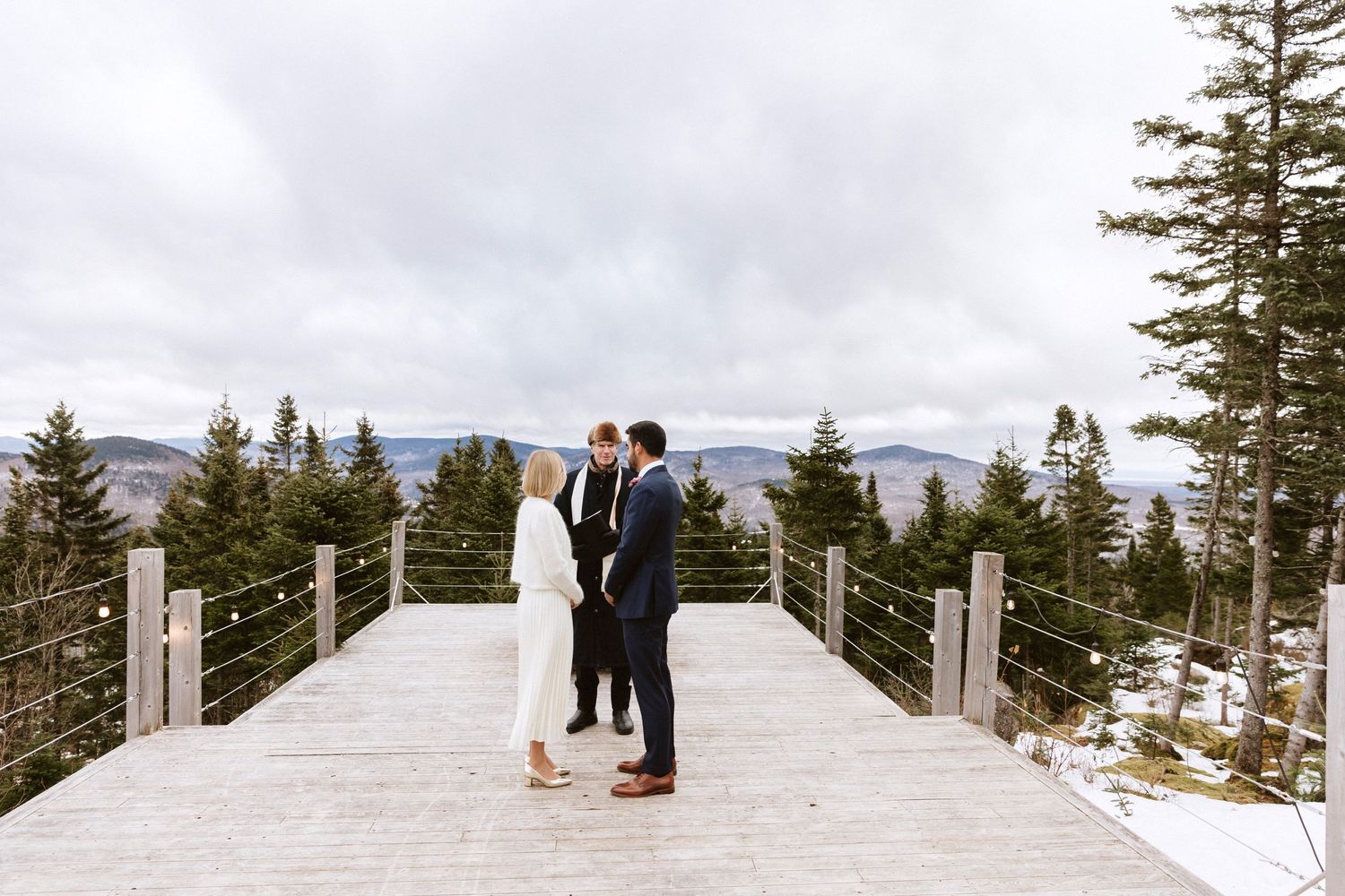 Winter wedding couple on an elevated wooden deck with mountain views and snow-covered trees.