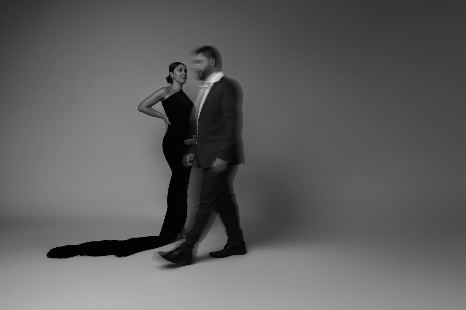 Elegant couple in formal attire poses dramatically in black and white studio setting.