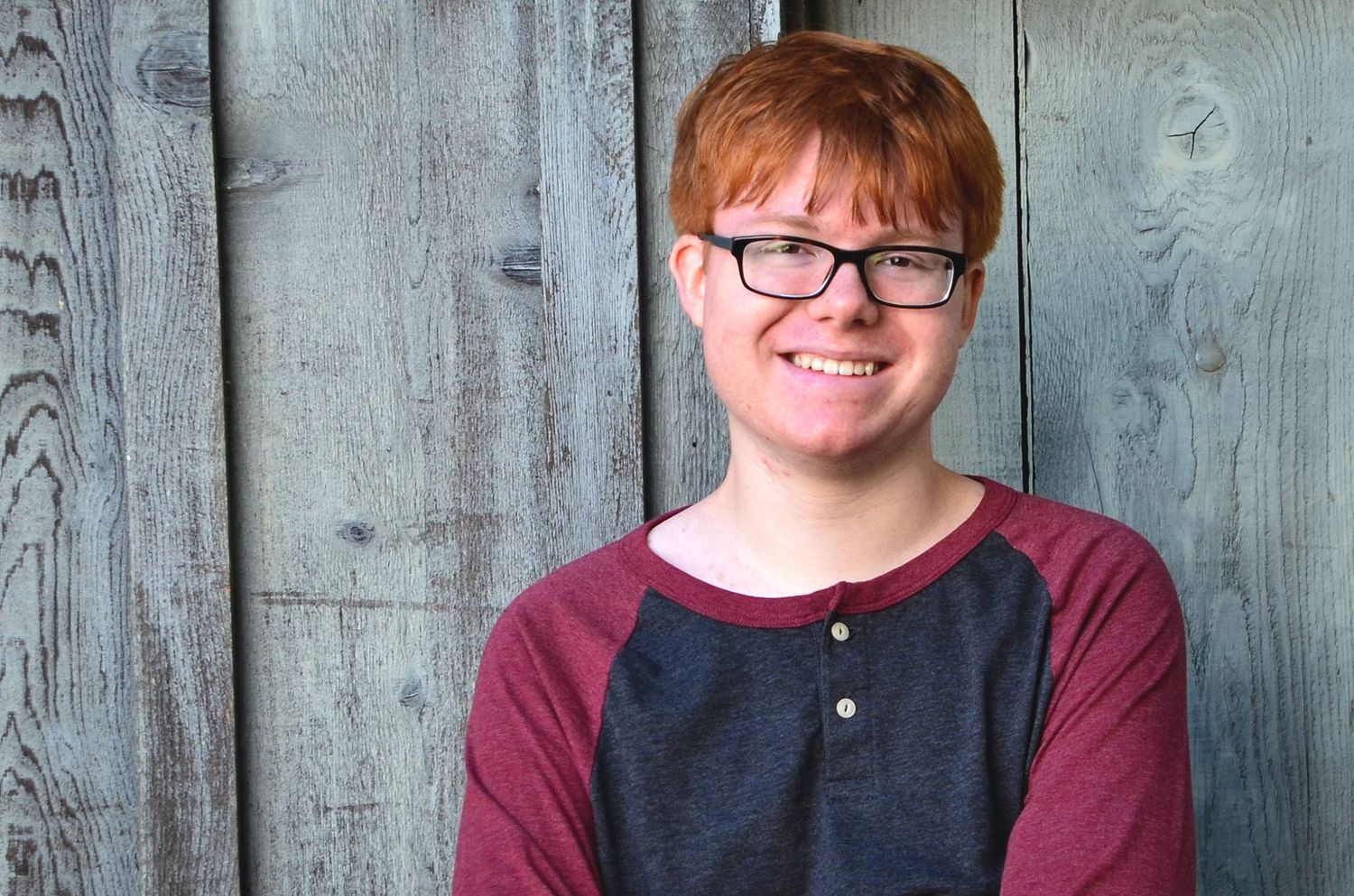 A teenager wearing a burgundy and grey baseball shirt poses against a wooden wall.