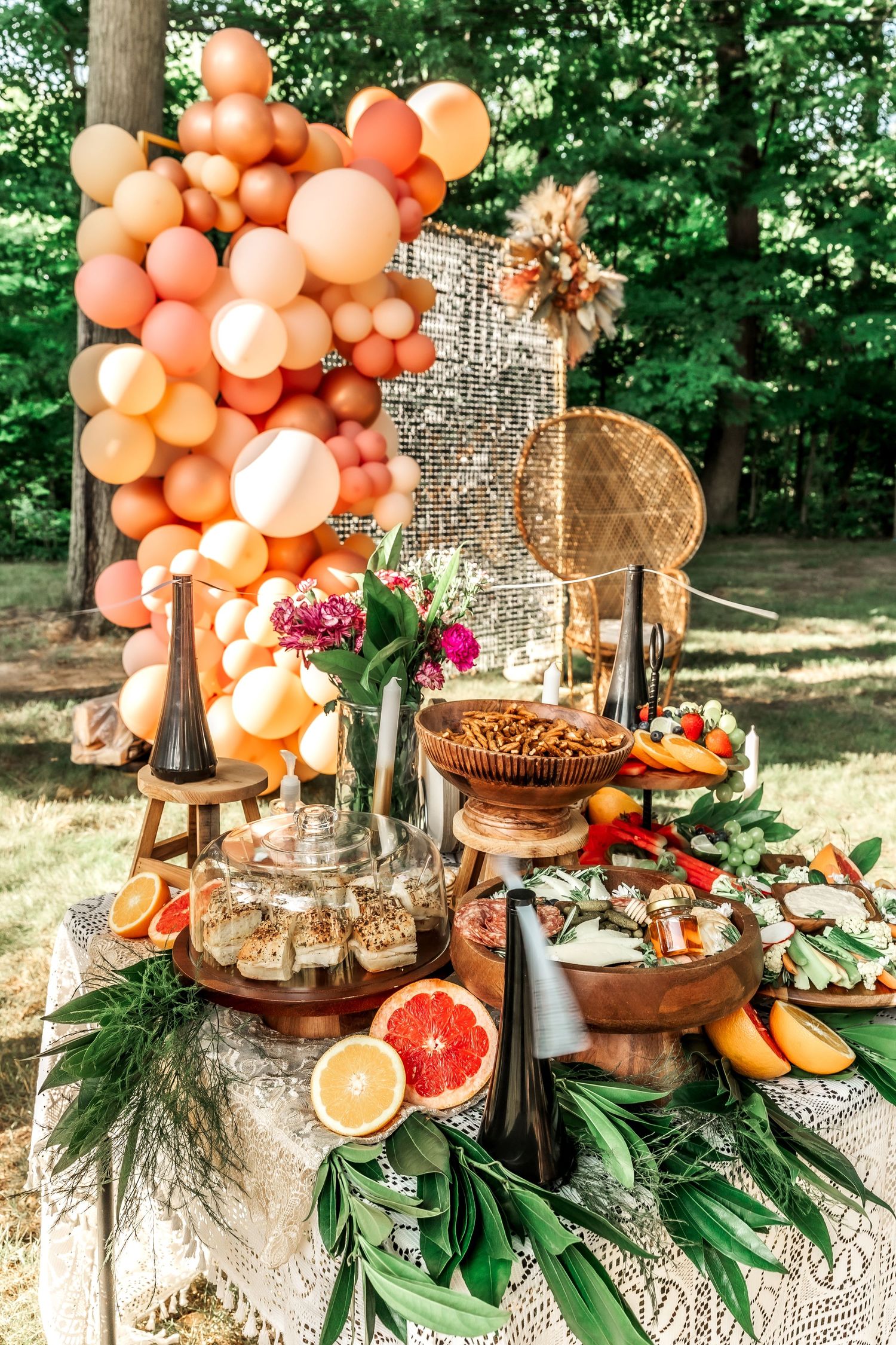 Elegant outdoor party setup with peach balloon arch, crystal decor, and grazing platters.