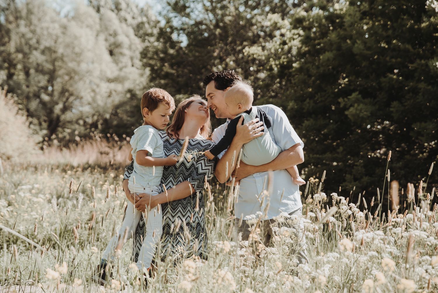 Family bonding moment captured in a sunny field with tall grass and trees.