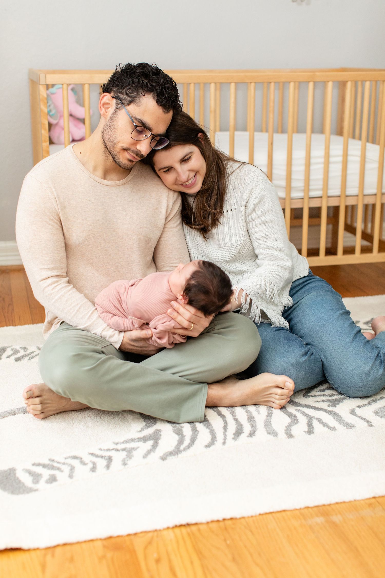 A couple sits on a blanket near a wooden crib while holding their sleeping newborn baby.