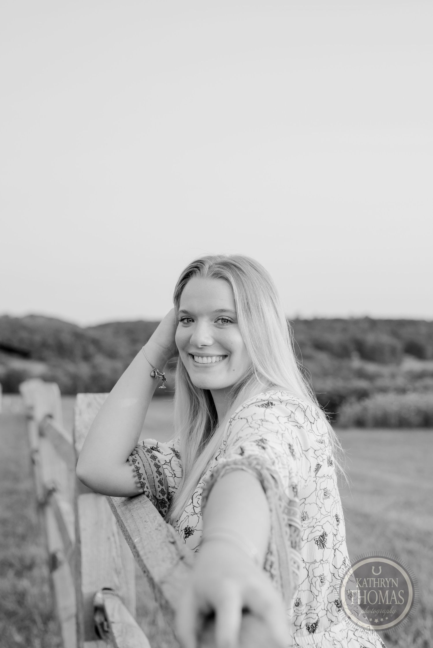Black and white portrait of a woman leaning on a rustic fence in a floral dress.