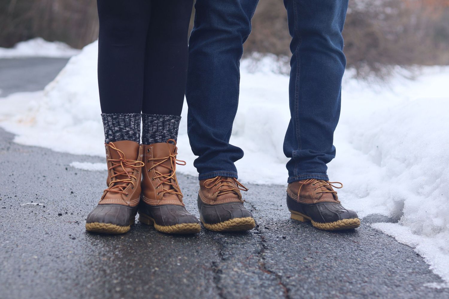 Two people stand side by side wearing matching L.L. Bean duck boots on a wet road with snow in the background.