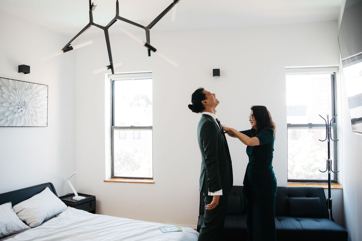 Couple in formal wear adjusting clothing in a bright minimal bedroom setting.