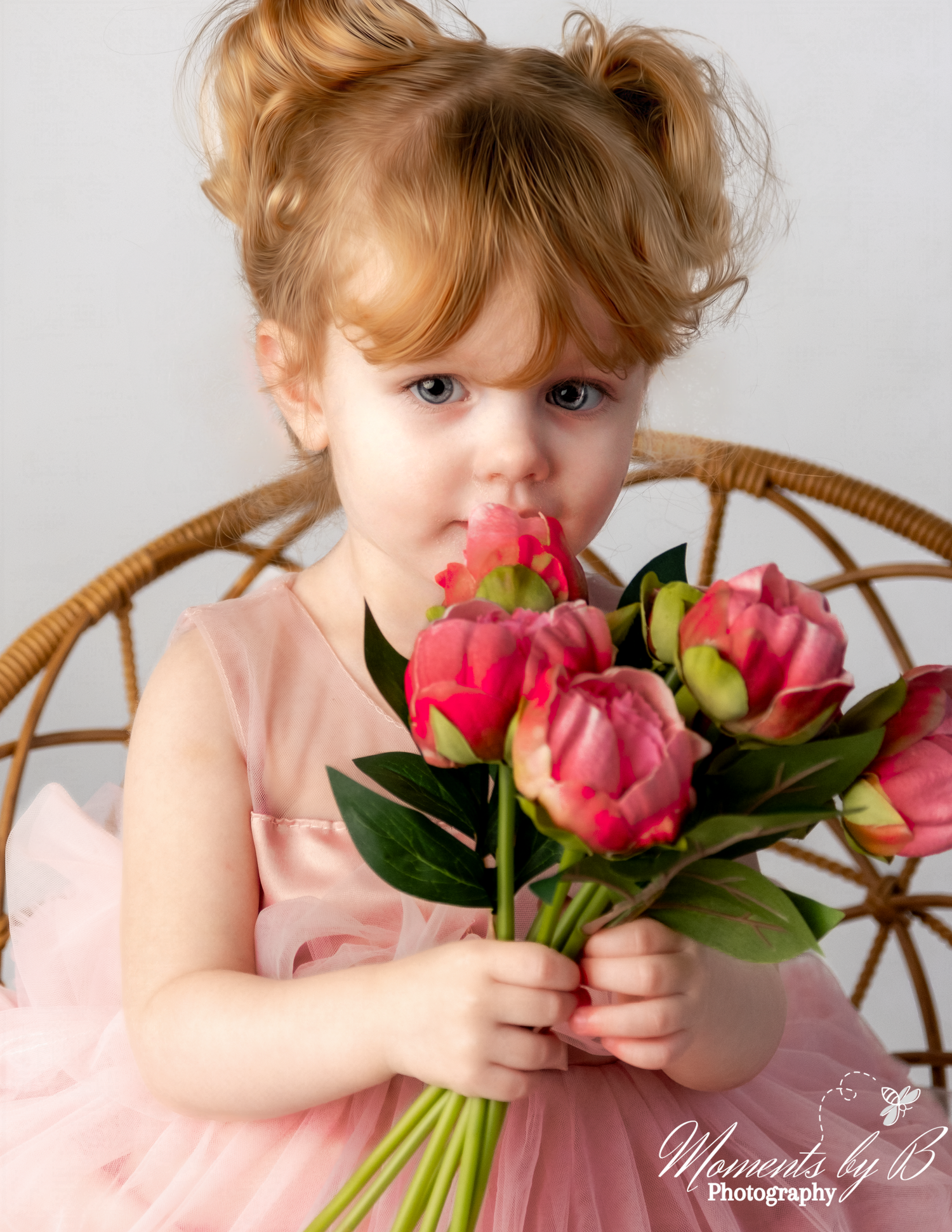 A small child in a pink dress holds a bouquet of vibrant pink tulips while sitting in a wicker chair.