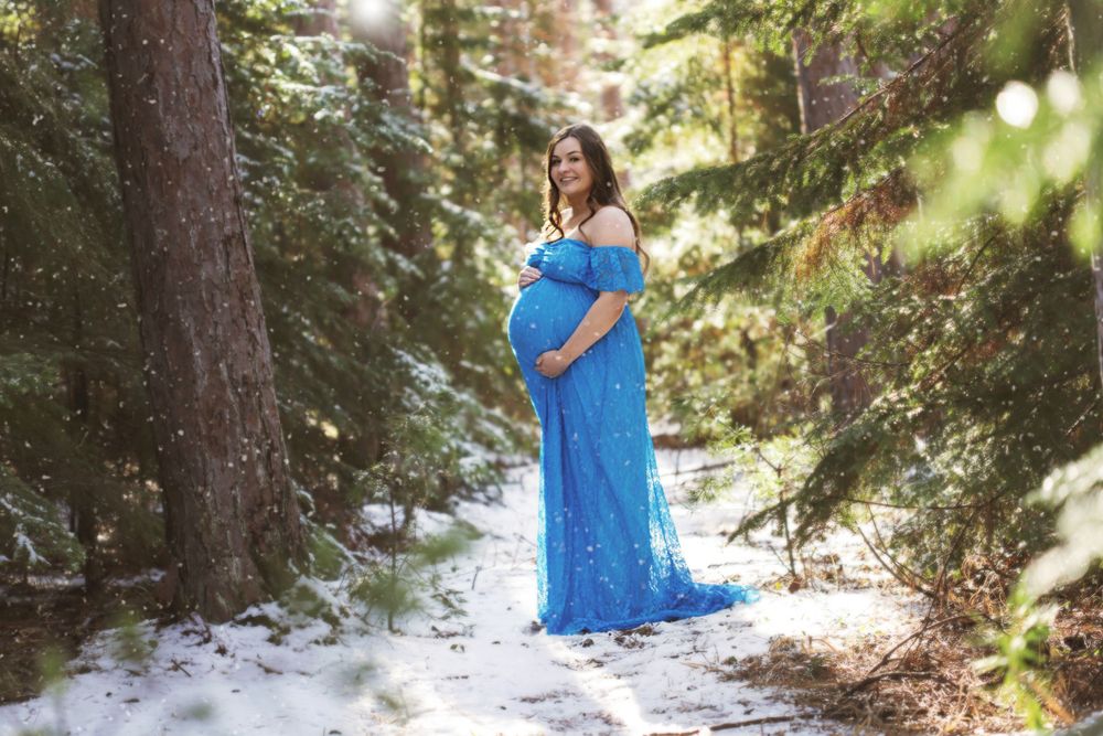 A pregnant woman in a flowing blue dress stands in a snowy forest setting during a maternity photoshoot. Duluth Minnesota Superior WI