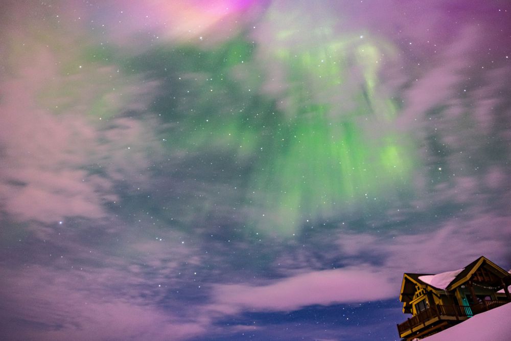 Vibrant purple and green aurora borealis dancing in night sky above cabin rooftop.
