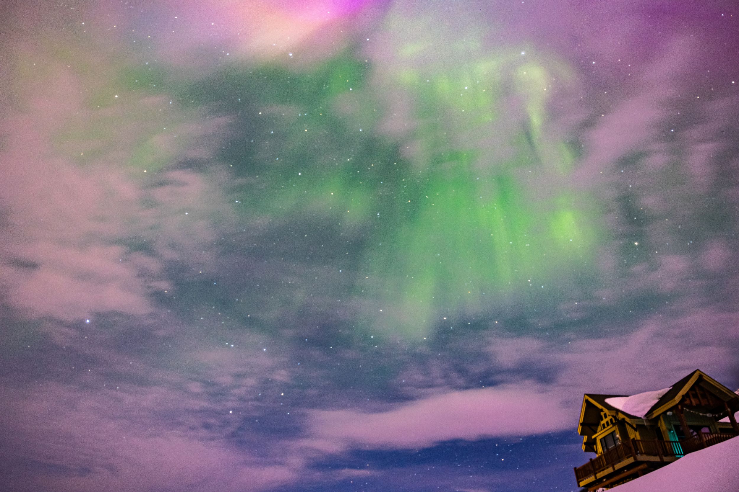 Vibrant purple and green aurora borealis dancing in night sky above cabin rooftop.