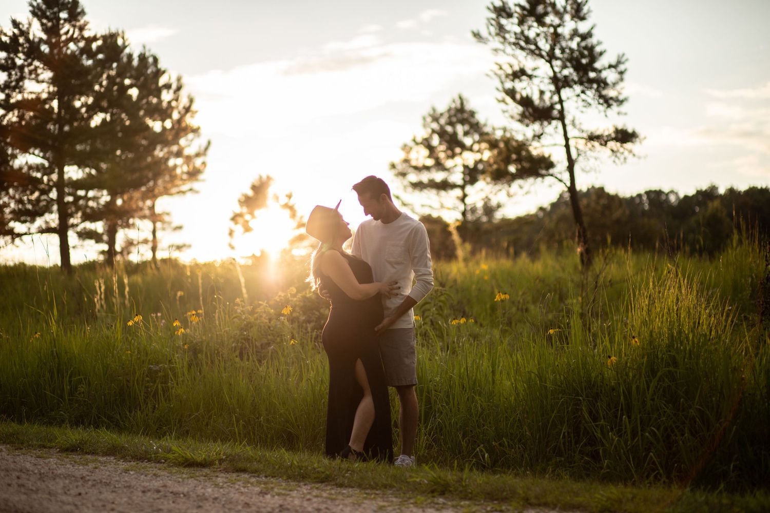Silhouetted couple embracing at sunset in grassy field with pine trees.