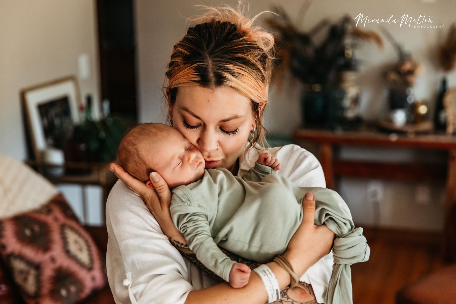 A mother holds her newborn baby and kisses him on the cheek. He wears a sage green infant gown.