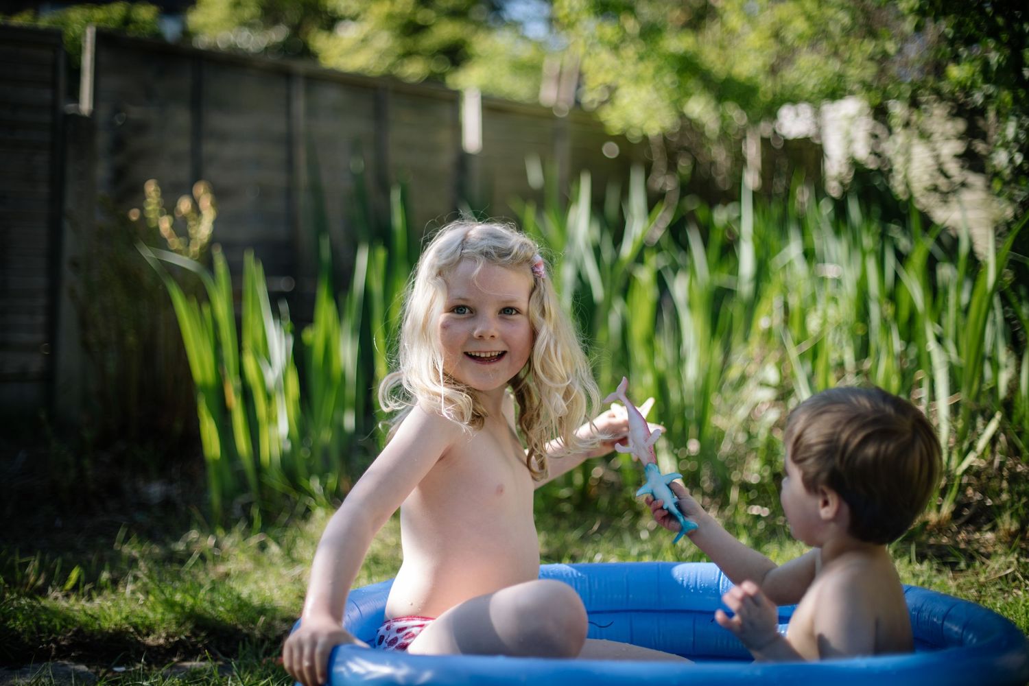 Two children playing together in a small blue inflatable pool in a backyard garden.