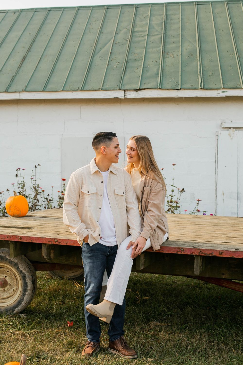 Couple in coordinating beige outfits posing by rustic barn with pumpkin decoration.