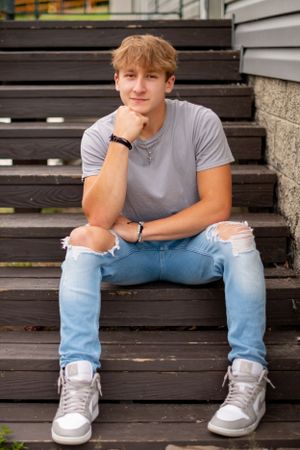 Young person in gray t-shirt and ripped jeans sitting casually on wooden steps.