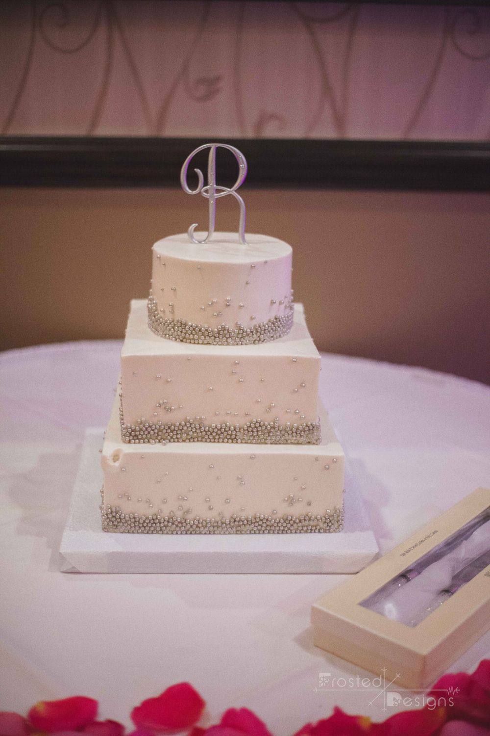 Wedding cake with letter 'R' on top with pink flowers on the table.