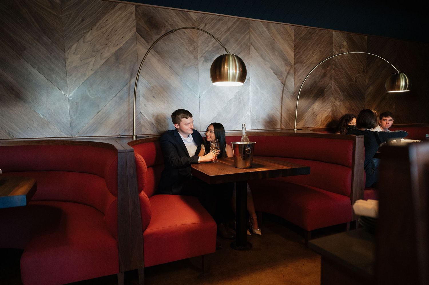 Romantic date night scene in dimly lit restaurant with curved red booths and modern pendant lighting.