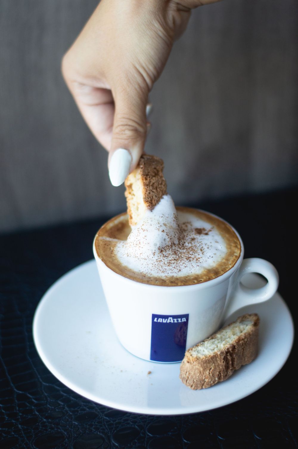 Hand sprinkling cocoa powder on a Lavazza cappuccino served with biscotti.