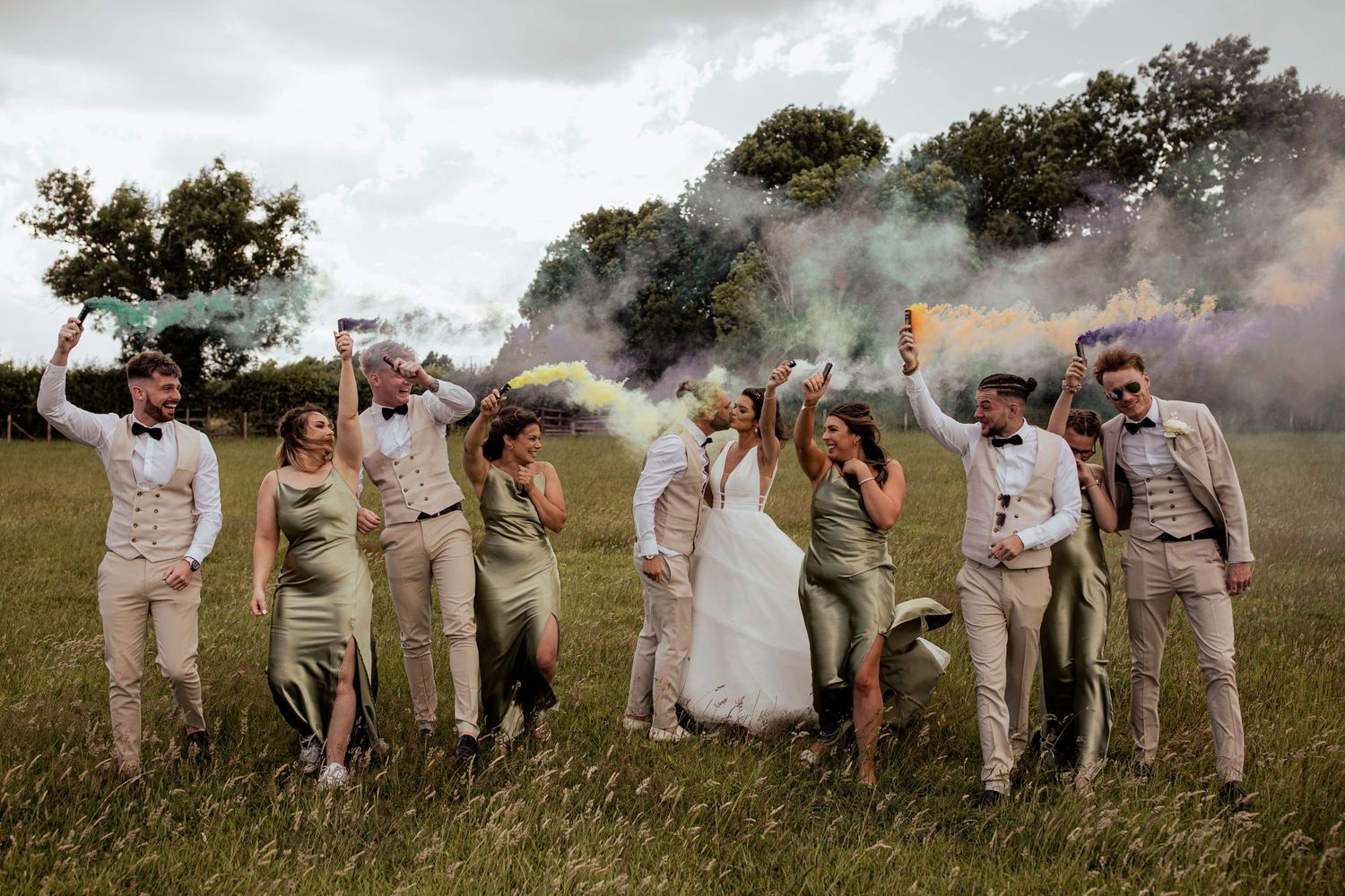Wedding party celebrates on a grassy field with colorful smoke bombs creating a dramatic effect.
