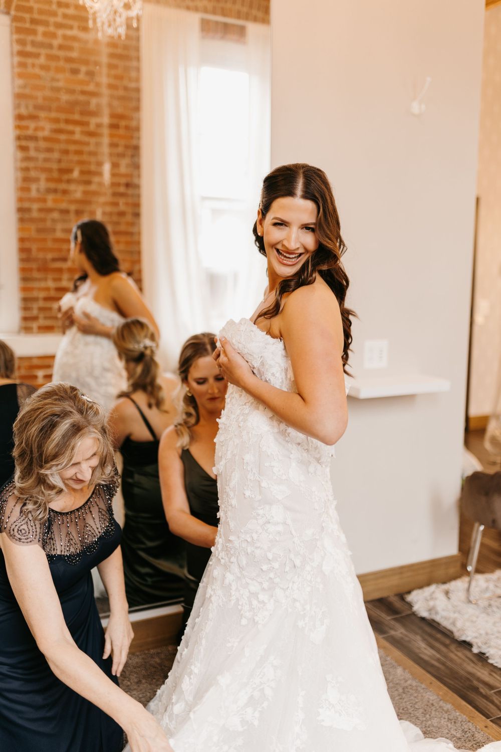 Radiant bride smiling while bridesmaids and family help her into her floral lace wedding dress.